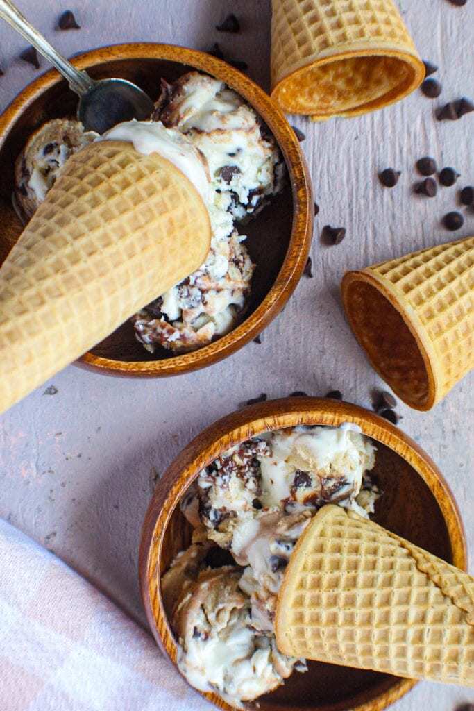 overhead shot of chocolate chip cookie dough ice cream in small wooden bowl with sugar cones placed on top of.
