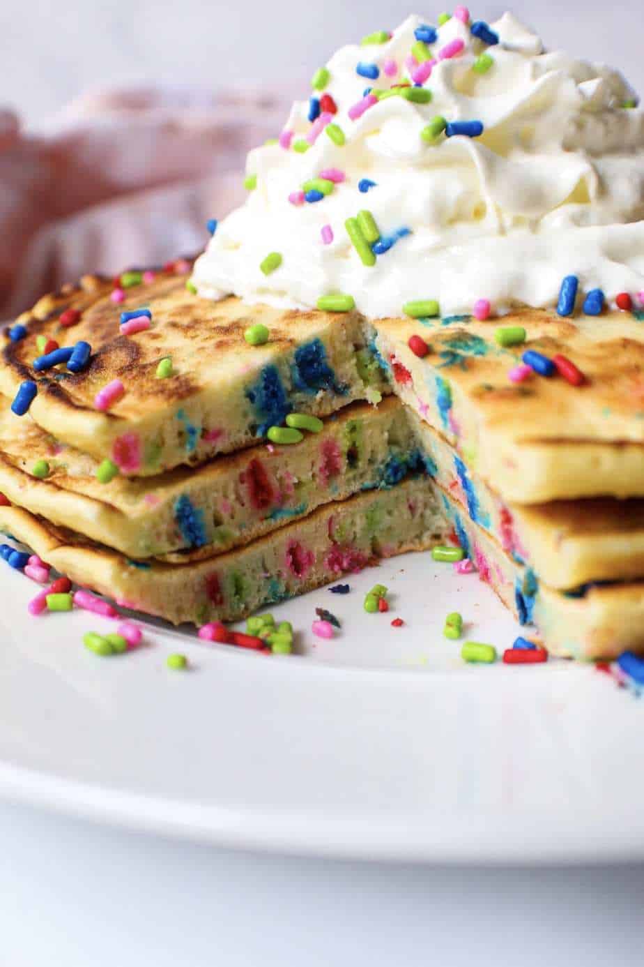 side angle shot of giant funfetti pancakes on a white plate with whipped cream on top.