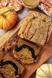 overhead shot of chocolate marble pumpkin bread on wooden board with orange floral towel and pumpkins in shot perimeter