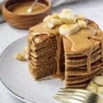 peanut butter banana pancakes in a stack with a section cut out topped with peanut butter drizzle and sliced bananas. bowl of peanut butter in background