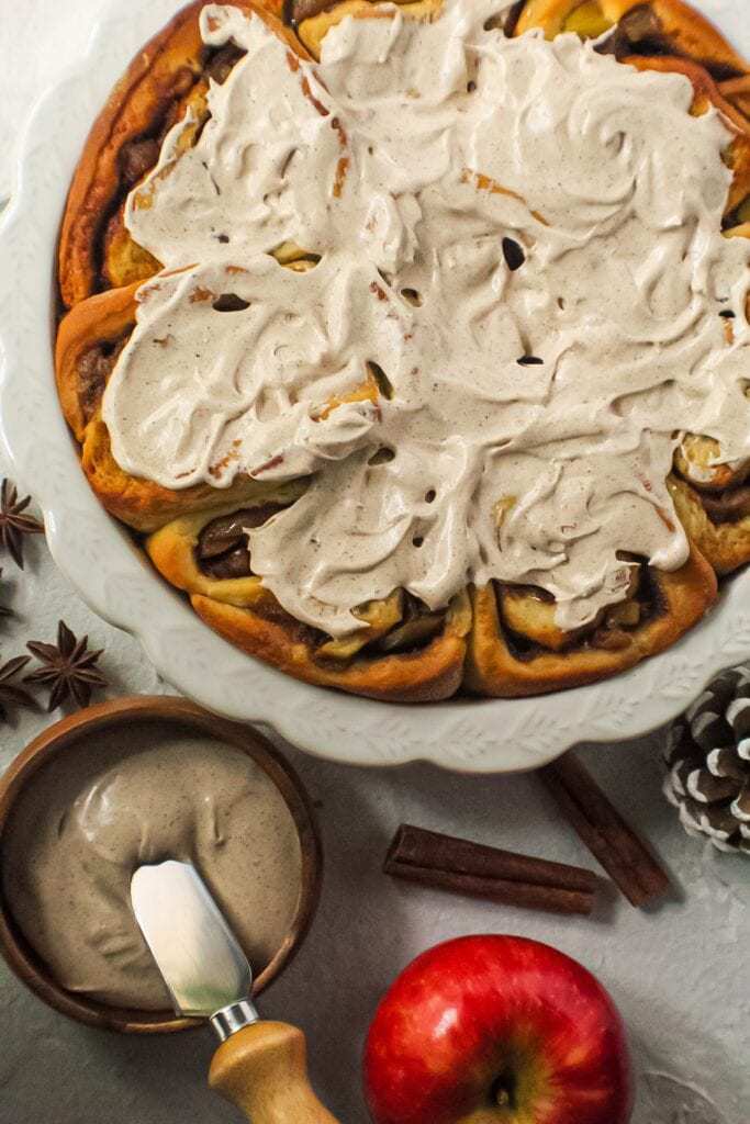 apple cinnamon rolls with chai cream cheese frosting on top in white pie dish with one cinnamon roll on a blue and white plate next to pie dish with cinnamon sticks and star anise in shot.