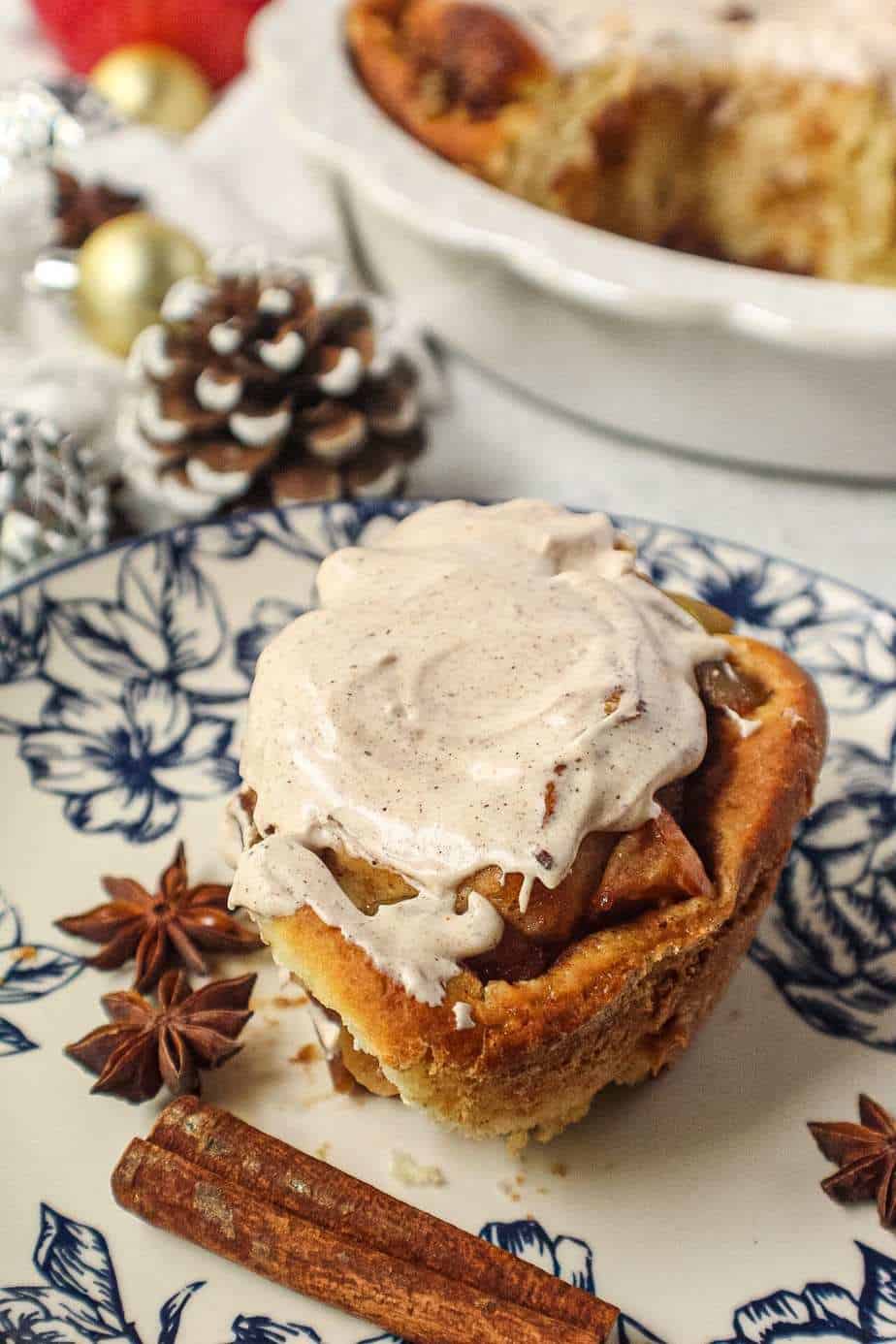 apple cinnamon rolls with chai cream cheese frosting on top in white pie dish with one cinnamon roll on a blue and white plate next to pie dish with cinnamon sticks and star anise in shot.