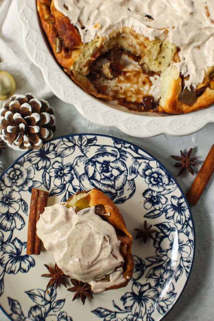 apple cinnamon rolls with chai cream cheese frosting on top in white pie dish with one cinnamon roll on a blue and white plate next to pie dish with cinnamon sticks and star anise in shot.