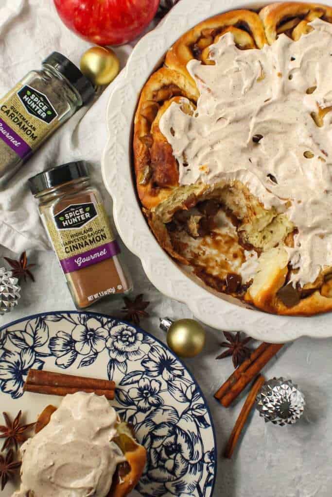 apple cinnamon rolls with chai cream cheese frosting on top in white pie dish with one cinnamon roll on a blue and white plate next to pie dish with cinnamon sticks and star anise in shot.