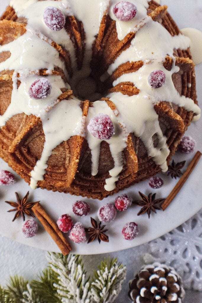 chai spice cake with maple cream cheese glaze on white serving plate with sugared cranberries on top and cinnamon sticks and star anise arranged around cake