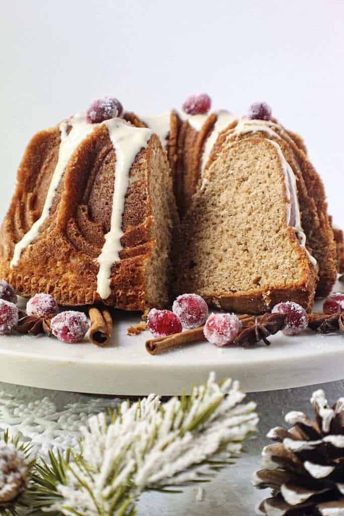 chai spice cake with maple cream cheese glaze on white serving plate with sugared cranberries on top and cinnamon sticks and star anise arranged around cake