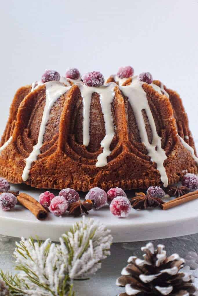 chai spice cake with maple cream cheese glaze on white serving plate with sugared cranberries on top and cinnamon sticks and star anise arranged around cake