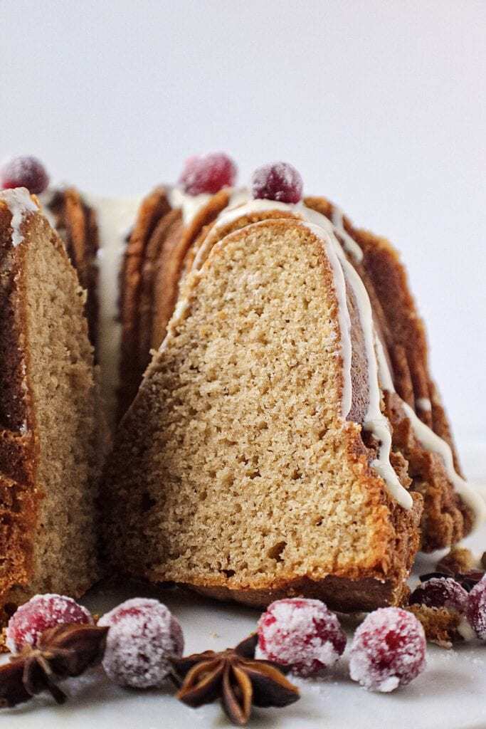 chai spice cake with maple cream cheese glaze on white serving plate with sugared cranberries on top and cinnamon sticks and star anise arranged around cake