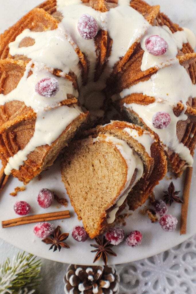 chai spice cake with maple cream cheese glaze on white serving plate with sugared cranberries on top and cinnamon sticks and star anise arranged around cake