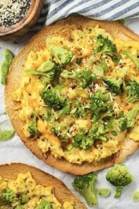 overhead shot of broccoli and cheese breakfast pizzas garnished with everything bagel seasoning on top of white wooden surface with striped linen in corner.