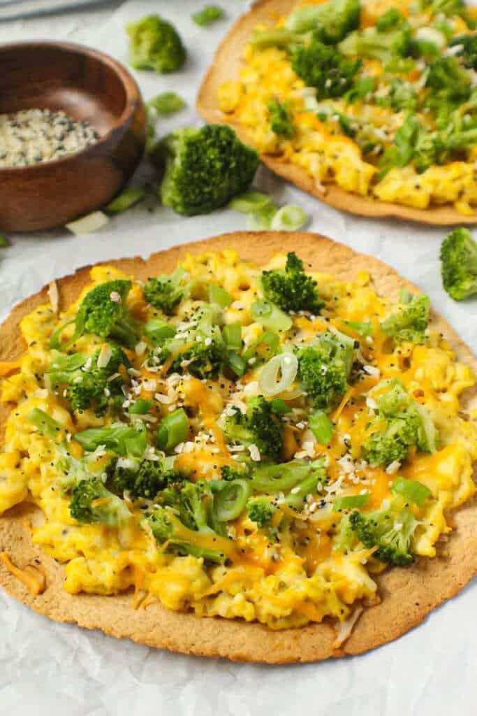 overhead shot of broccoli and cheese breakfast pizzas garnished with everything bagel seasoning on top of white wooden surface with striped linen in corner.