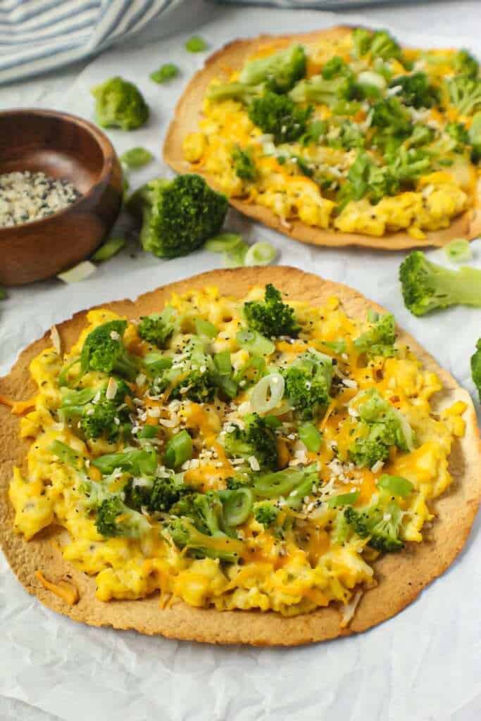 overhead shot of broccoli and cheese breakfast pizzas garnished with everything bagel seasoning on top of white wooden surface with striped linen in corner.