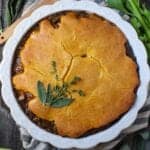 Cornbread Vegetable Pot Pie in a glass pie dish garnished with fresh herbs on top of striped linen on wood surface with a wooden spoon and vegetables around the pie dish