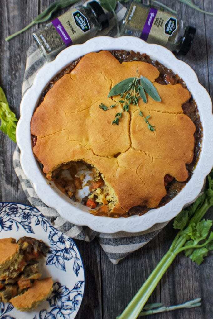 Cornbread Vegetable Pot Pie in a glass pie dish garnished with fresh herbs on top of striped linen on wood surface with a wooden spoon, spice bottles and vegetables around the pie dish