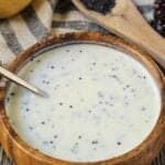Creamy Lemon Poppyseed Dressing in a wooden bowl with wooden spoon filled with poppy seeds in shot along with a whole lemon, leaf lettuce and cloth napkin