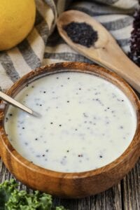 Creamy Lemon Poppyseed Dressing in a wooden bowl with wooden spoon filled with poppy seeds in shot along with a whole lemon, leaf lettuce and cloth napkin