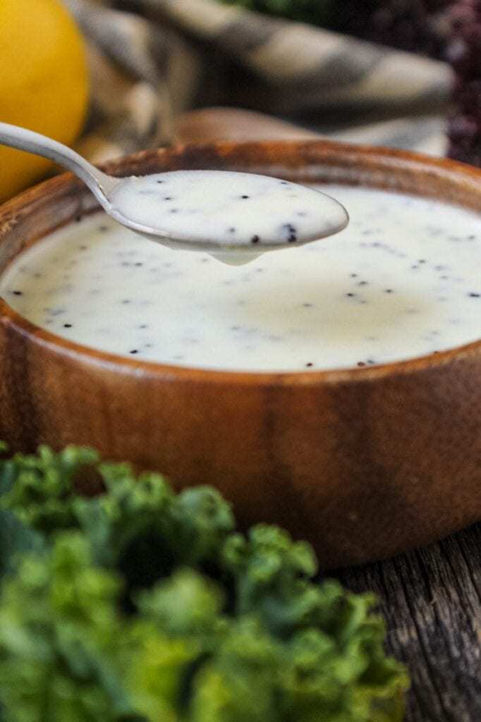 Creamy Lemon Poppyseed Dressing in a wooden bowl with wooden spoon filled with poppy seeds in shot along with a whole lemon, leaf lettuce and cloth napkin