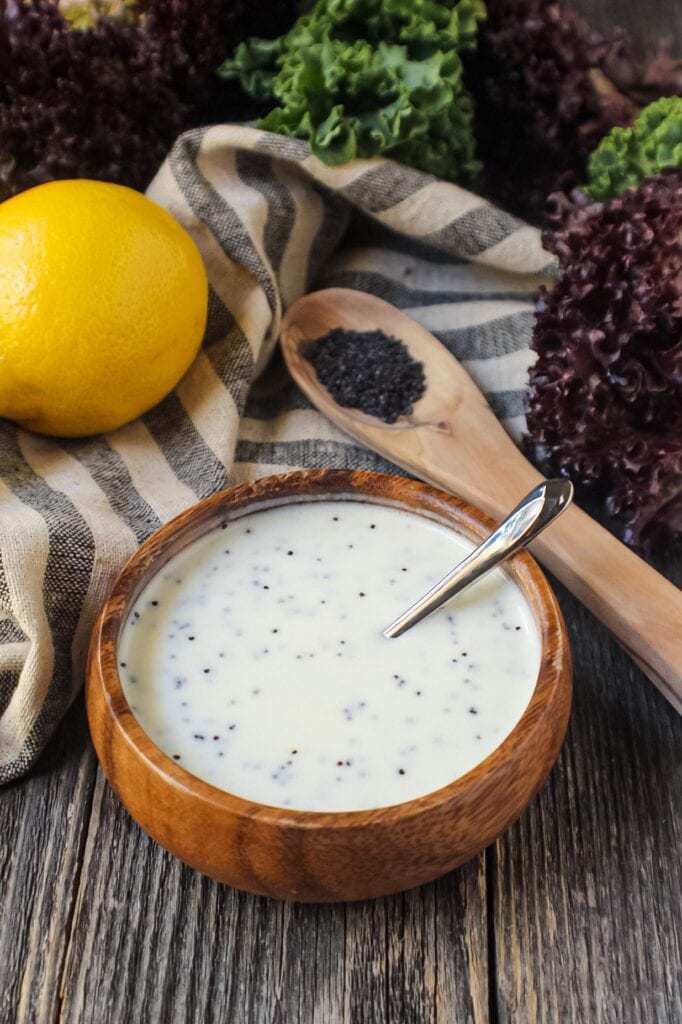 Creamy Lemon Poppyseed Dressing in a wooden bowl with wooden spoon filled with poppy seeds in shot along with a whole lemon, leaf lettuce and cloth napkin