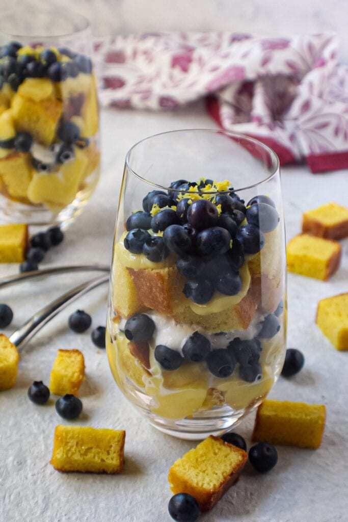 side shot of lemon blueberry trifles served in wine glasses with cake pieces scattered on surface and colorful linen in background