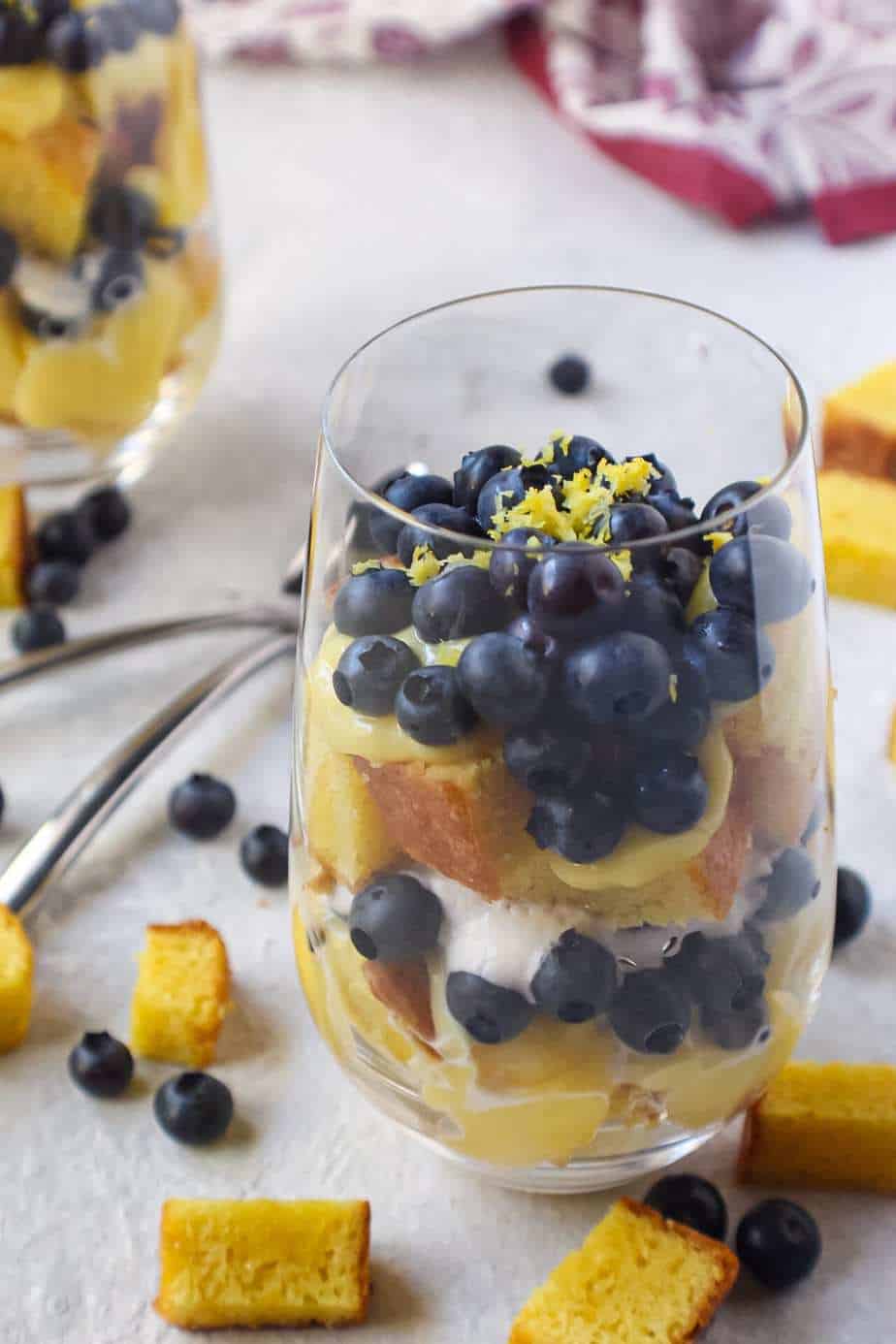side shot of lemon blueberry trifles served in wine glasses with cake pieces scattered on surface and colorful linen in background
