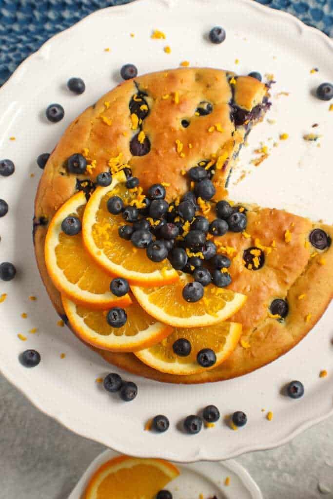 orange blueberry breakfast cake on white cake stand with fresh blueberries on top and garnished with fresh orange slices and orange zest. Half an orange and blue towel in the background.