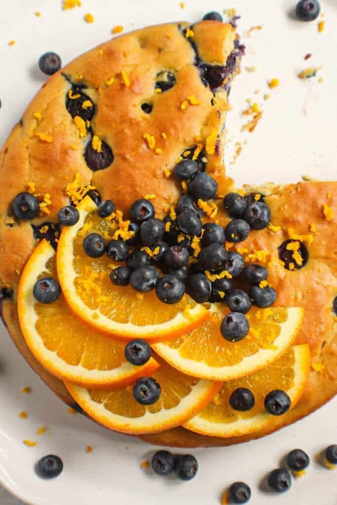 overhead shot of orange blueberry breakfast cake on white cake stand with fresh blueberries on top and garnished with fresh orange slices and orange zest. Half an orange and blue towel in the background.
