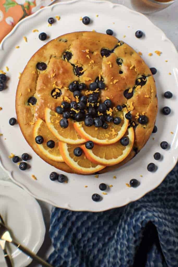 overhead shot of orange blueberry breakfast cake on white cake stand with fresh blueberries on top and garnished with fresh orange slices and orange zest. Half an orange and blue towel in the background.y breakfast cake