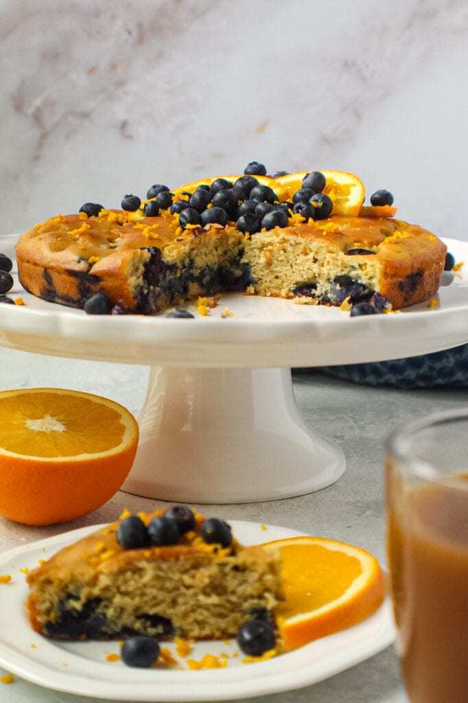 side angle shot of orange blueberry breakfast cake on white cake stand with fresh blueberries on top and garnished with fresh orange slices and orange zest. Half an orange and blue towel in the background.y breakfast cake