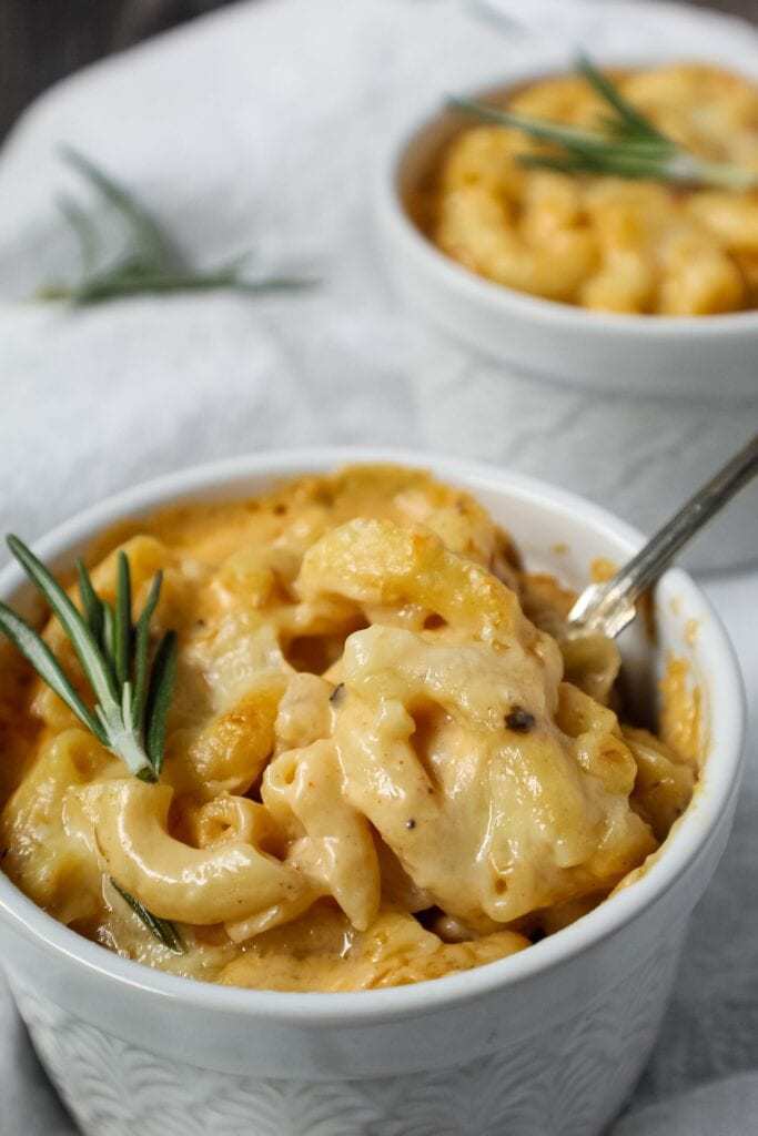 smoked cheddar mac and cheese served in two single-serve ramekins sitting on top of white linen topped with fresh rosemary and a small spoon beside ramekin.