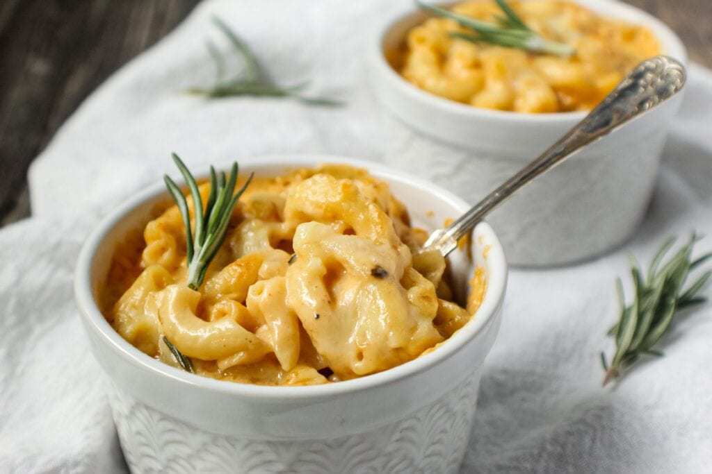 smoked cheddar mac and cheese served in two single-serve ramekins sitting on top of white linen topped with fresh rosemary and a small spoon beside ramekin.