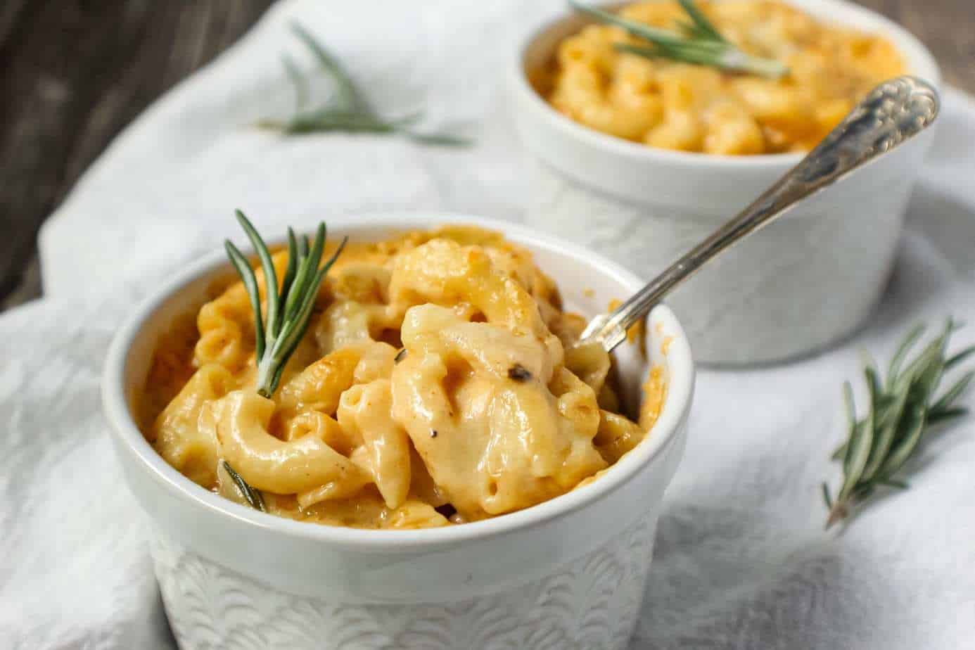 smoked cheddar mac and cheese served in two single-serve ramekins sitting on top of white linen topped with fresh rosemary and a small spoon beside ramekin.
