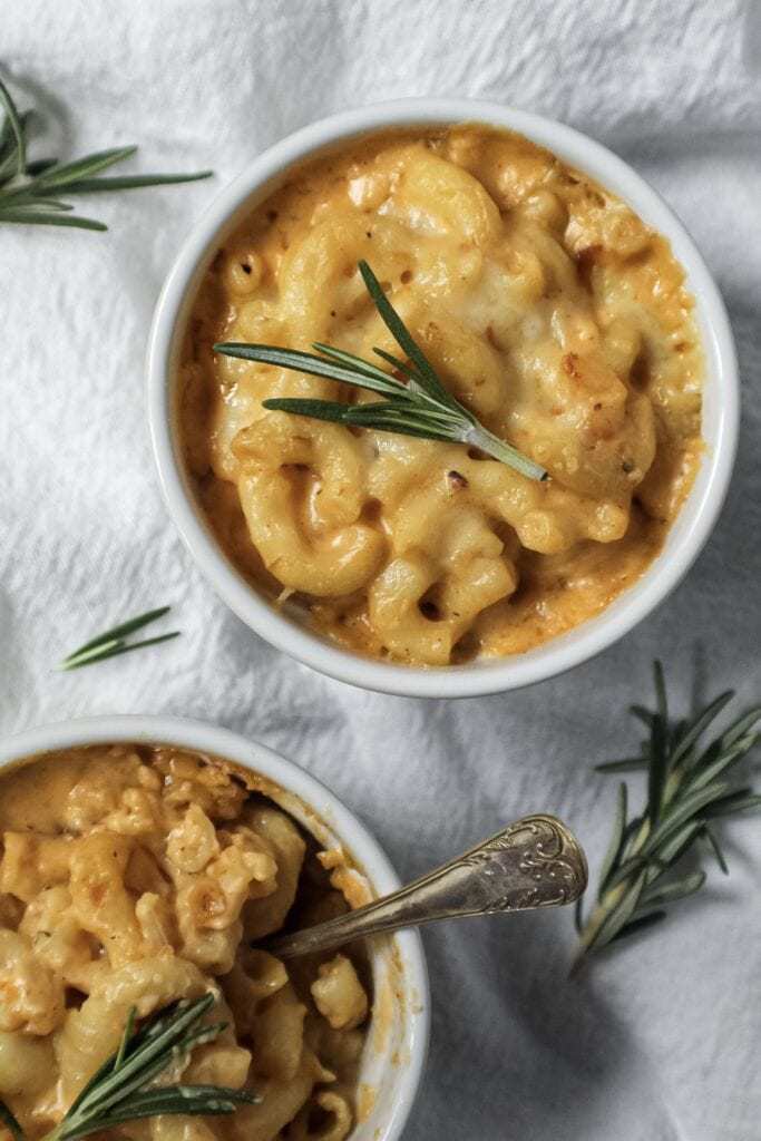 smoked cheddar mac and cheese served in two single-serve ramekins sitting on top of white linen topped with fresh rosemary and a small spoon inside one ramekin.