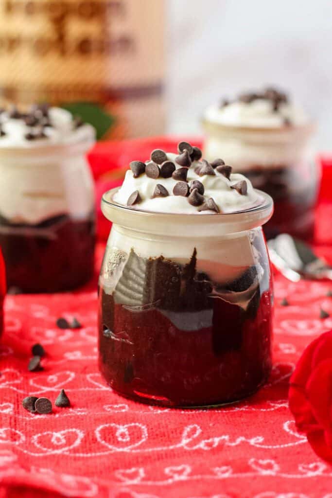 Dark chocolate avocado mousse in a yogurt jar topped with dairy-free yogurt and mini chocolate chips. Jars are on top of a red kitchen towel.