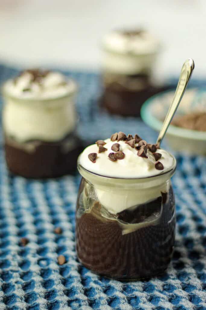 Dark chocolate avocado mousse in a yogurt jar topped with dairy-free yogurt and mini chocolate chips. Jars are on top of a blue kitchen towel.