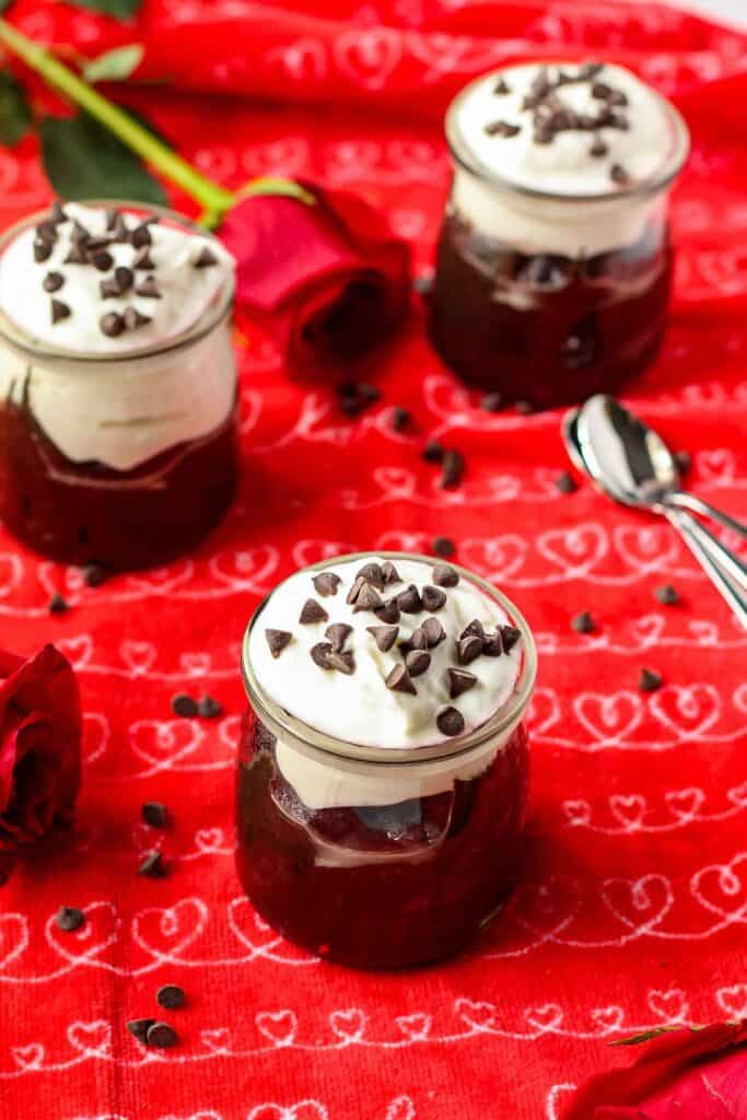 Dark chocolate avocado mousse in a yogurt jar topped with dairy-free yogurt and mini chocolate chips. Jars are on top of a red kitchen towel.