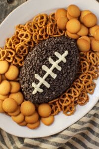 Cookies and cream cheese ball shaped and decorated to look like a football. sitting on a white serving platter with pretzels and vanilla wafer cookies around cheese ball.