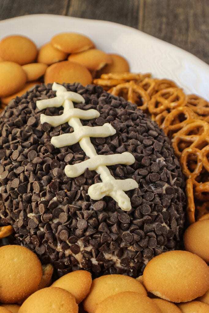 Cookies and cream cheese ball shaped and decorated to look like a football. sitting on a white serving platter with pretzels and vanilla wafer cookies around cheese ball.