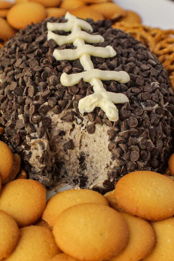 Cookies and cream cheese ball shaped and decorated to look like a football. sitting on a white serving platter with pretzels and vanilla wafer cookies around cheese ball.