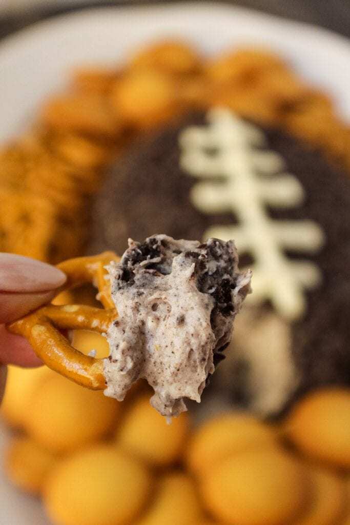 Cookies and cream cheese ball shaped and decorated to look like a football. sitting on a white serving platter with pretzels and vanilla wafer cookies around cheese ball in background. pretzel with cheese ball filling on in in forefront of shot