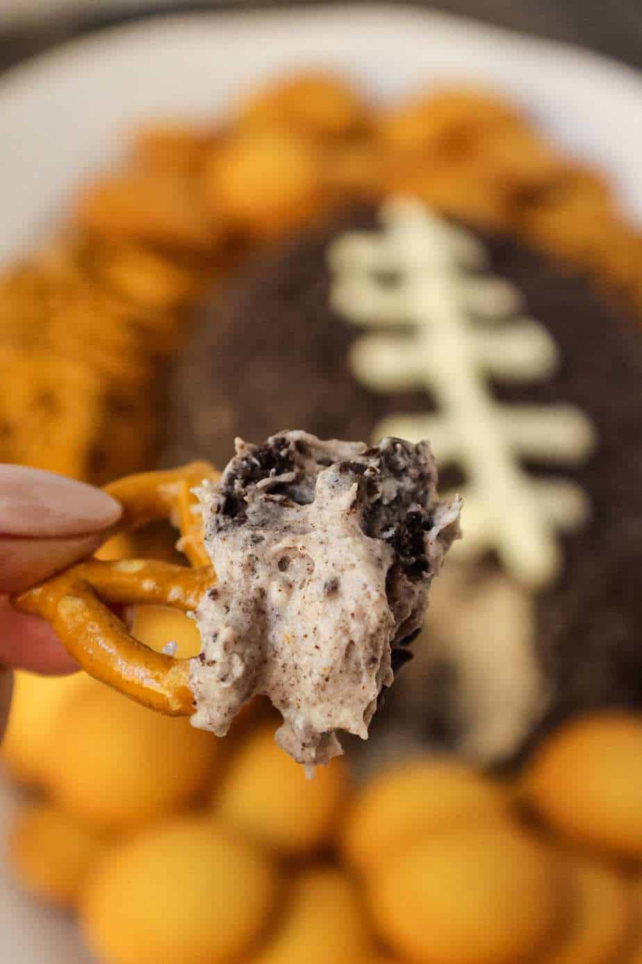 Cookies and cream cheese ball shaped and decorated to look like a football. sitting on a white serving platter with pretzels and vanilla wafer cookies around cheese ball in background. pretzel with cheese ball filling on in in forefront of shot