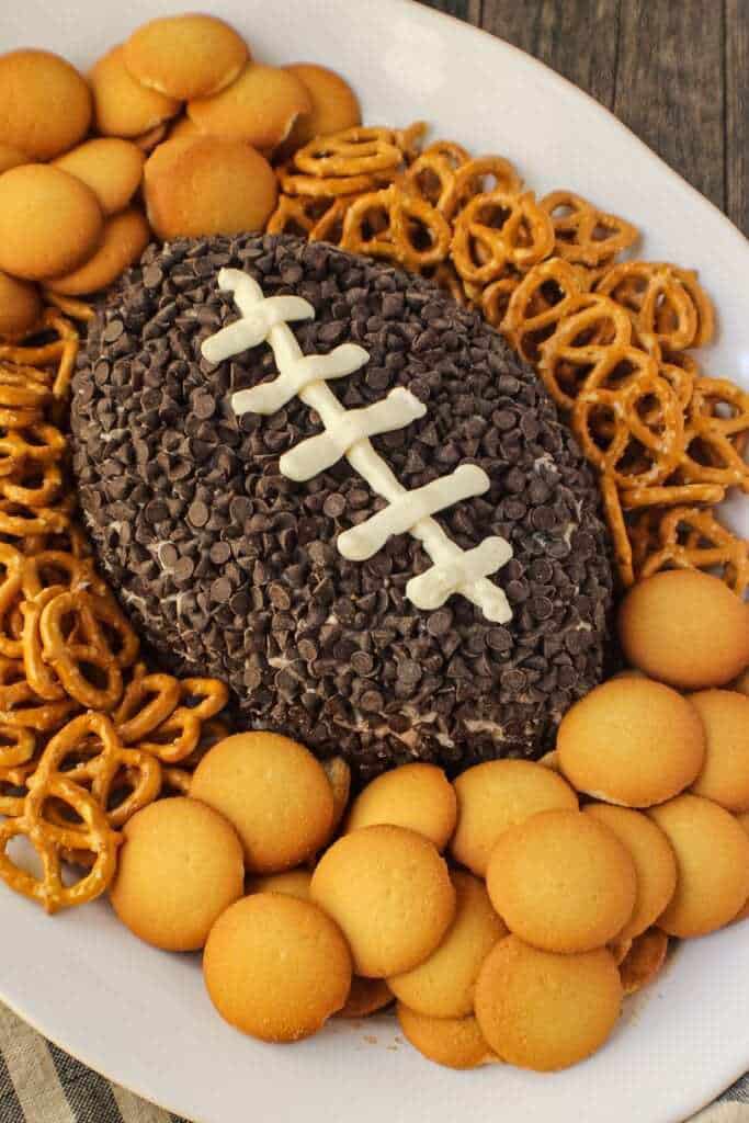 Cookies and cream cheese ball shaped and decorated to look like a football. sitting on a white serving platter with pretzels and vanilla wafer cookies around cheese ball.