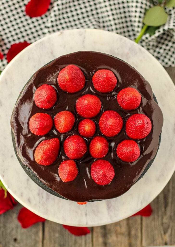overhead shot of dark chocolate strawberry cake with chocolate ganache on top of marble and wood cake stand with strawberries on top of cake and cloth napkin in background/