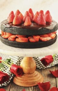 side angle shot of dark chocolate strawberry cake with chocolate ganache on top of marble and wood cake stand with strawberries arranged on top of cake and roses and cloth napkin in background.