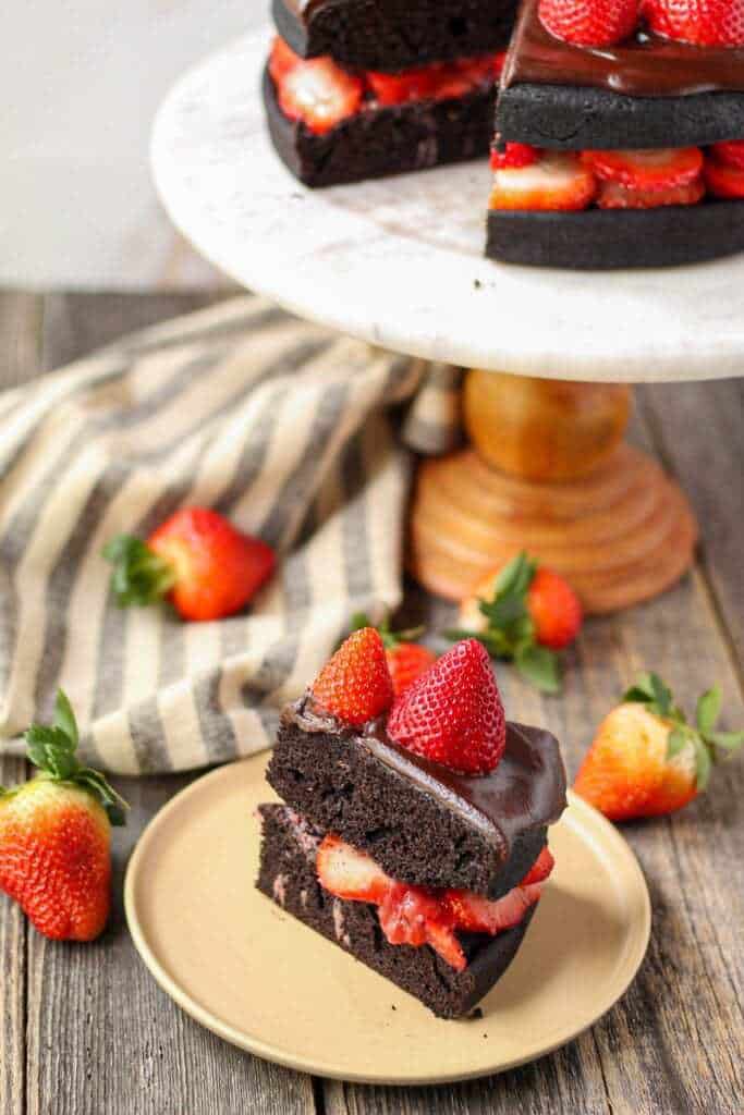 side angle shot of dark chocolate strawberry cake with chocolate ganache on top of marble and wood cake stand with a slice cut from the cake placed on small plate in bottom of shot.