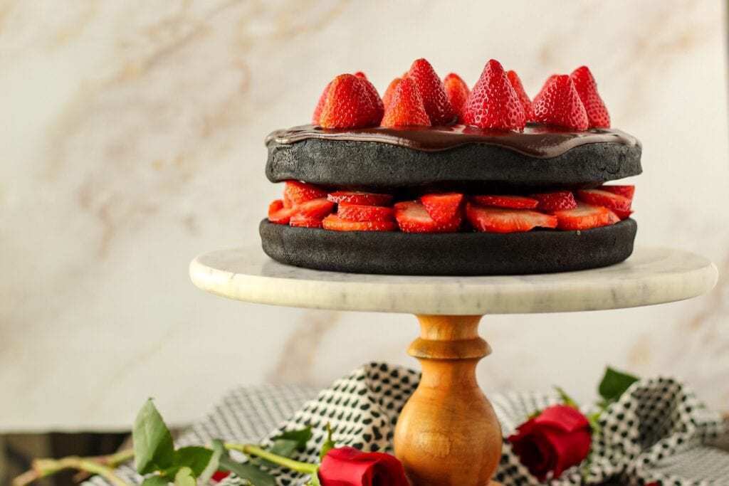 side angle shot of dark chocolate strawberry cake with chocolate ganache on top of marble and wood cake stand with cloth napkin and roses in background.