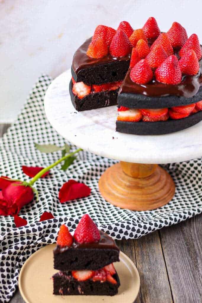 side angle shot of dark chocolate strawberry cake with chocolate ganache on top of marble and wood cake stand with a slice cut from the cake placed on small plate in bottom of shot.
