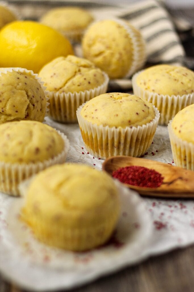 side angle shot of Lemon Cranberry Seed Cupcakes with Lemon Greek Yogurt Frosting on top parchment paper with cranberry seeds scattered on parchment and a wooden spoon filled with cranberry seeds in bottom right of shot. cloth napkin and whole lemon in background.