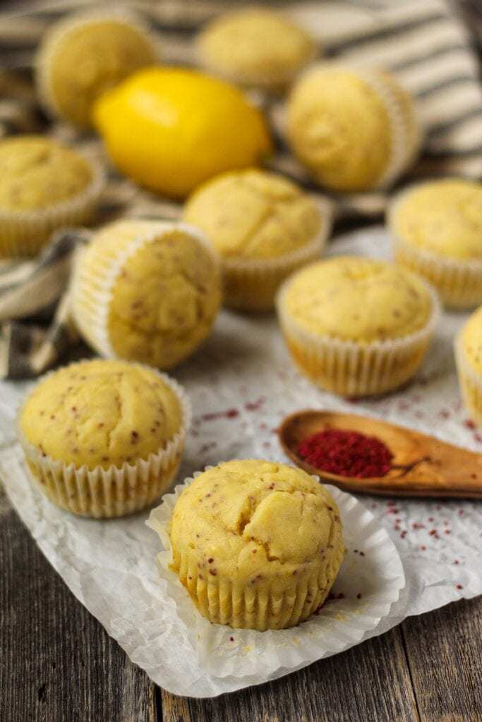 side angle shot of Lemon Cranberry Seed Cupcakes with Lemon Greek Yogurt Frosting on top parchment paper with cranberry seeds scattered on parchment and a wooden spoon filled with cranberry seeds in bottom right of shot. Cloth napkin and whole lemon in background.