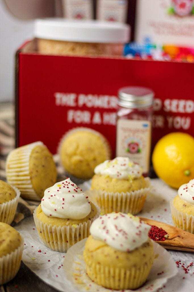 side angle shot of Lemon Cranberry Seed Cupcakes with Lemon Greek Yogurt Frosting on top parchment paper with cranberry seeds scattered on parchment and a wooden spoon filled with cranberry seeds in bottom right of shot. Cranberry seed container and box in background.