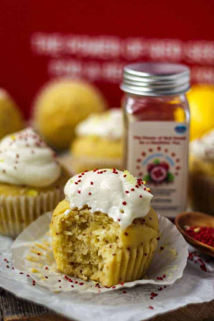 side angle shot of Lemon Cranberry Seed Cupcakes with Lemon Greek Yogurt Frosting on top parchment paper with cranberry seeds scattered on parchment and a wooden spoon filled with cranberry seeds in bottom right of shot. Cranberry seed container and box in background.